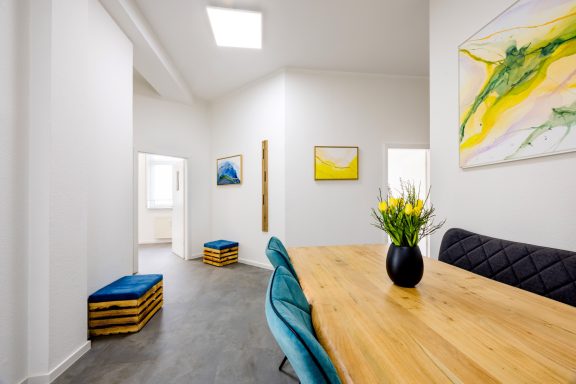 Heller Raum mit Holzstisch, blauen Stühlen und bunten Bildern an der Wand.